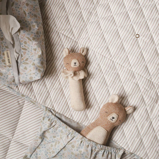 Knitted bear-themed baby rattle and soft toy resting on a quilted beige and striped baby blanket, surrounded by floral-patterned nursery accessories in a neutral-toned setting.