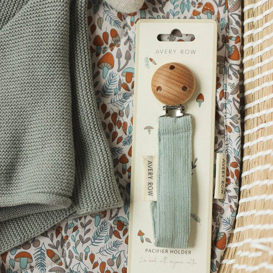 A Blue corduroy pacifier clip with a round wooden clasp at the top and a soft fabric strap leading to a silicone ring for attaching a pacifier. A small off-white tag on the side reads “AVERY ROW” in simple, elegant text. The minimalist design and muted color make it a stylish and practical accessory for keeping baby’s pacifier within reach.