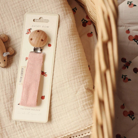 A blush pink corduroy pacifier clip with a round wooden clasp at the top and a soft fabric strap that loops into a silicone ring for attaching a pacifier. A small cream-colored label on the side reads “AVERY ROW” in simple lettering. The clip has a minimalist, elegant look, combining natural materials and gentle colors for a stylish and practical baby accessory.
