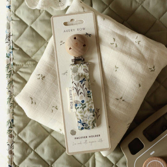 A stylish pacifier clip with a natural wood button clasp at the top and a fabric strap featuring a delicate botanical print in muted greens, blues, and browns. The strap is labeled “AVERY ROW” on a small fabric tag and ends with a silicone ring for attaching a pacifier. The design is both practical and charming, offering a secure and beautiful way to keep a baby’s pacifier within reach