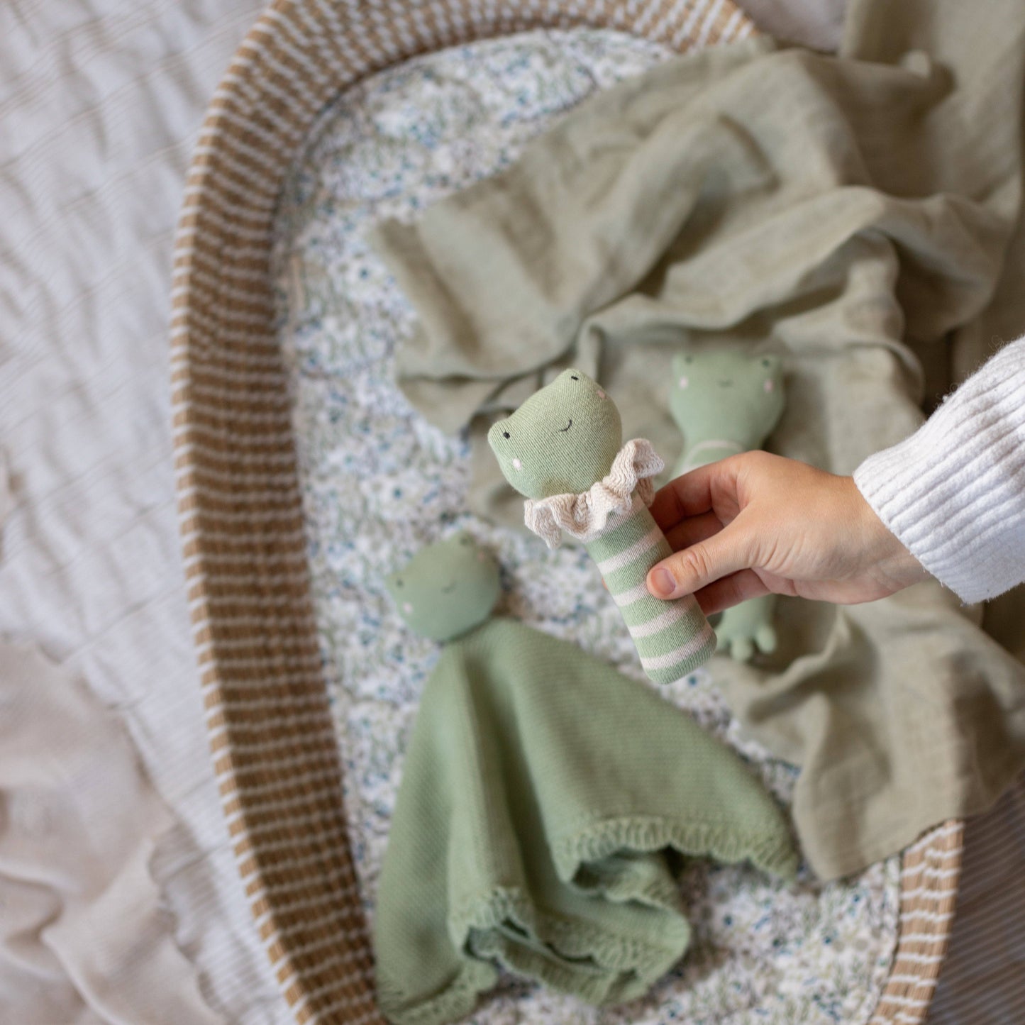 Hand holding a soft green frog-themed baby rattle with a white frill collar, surrounded by matching plush toys and a knitted comforter in a cozy, patterned baby bassinet.