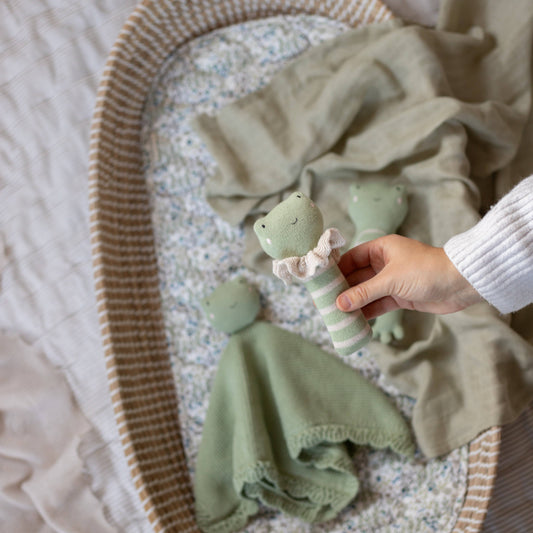 Hand holding a soft green frog-themed baby rattle with a white frill collar, surrounded by matching plush toys and a knitted comforter in a cozy, patterned baby bassinet.