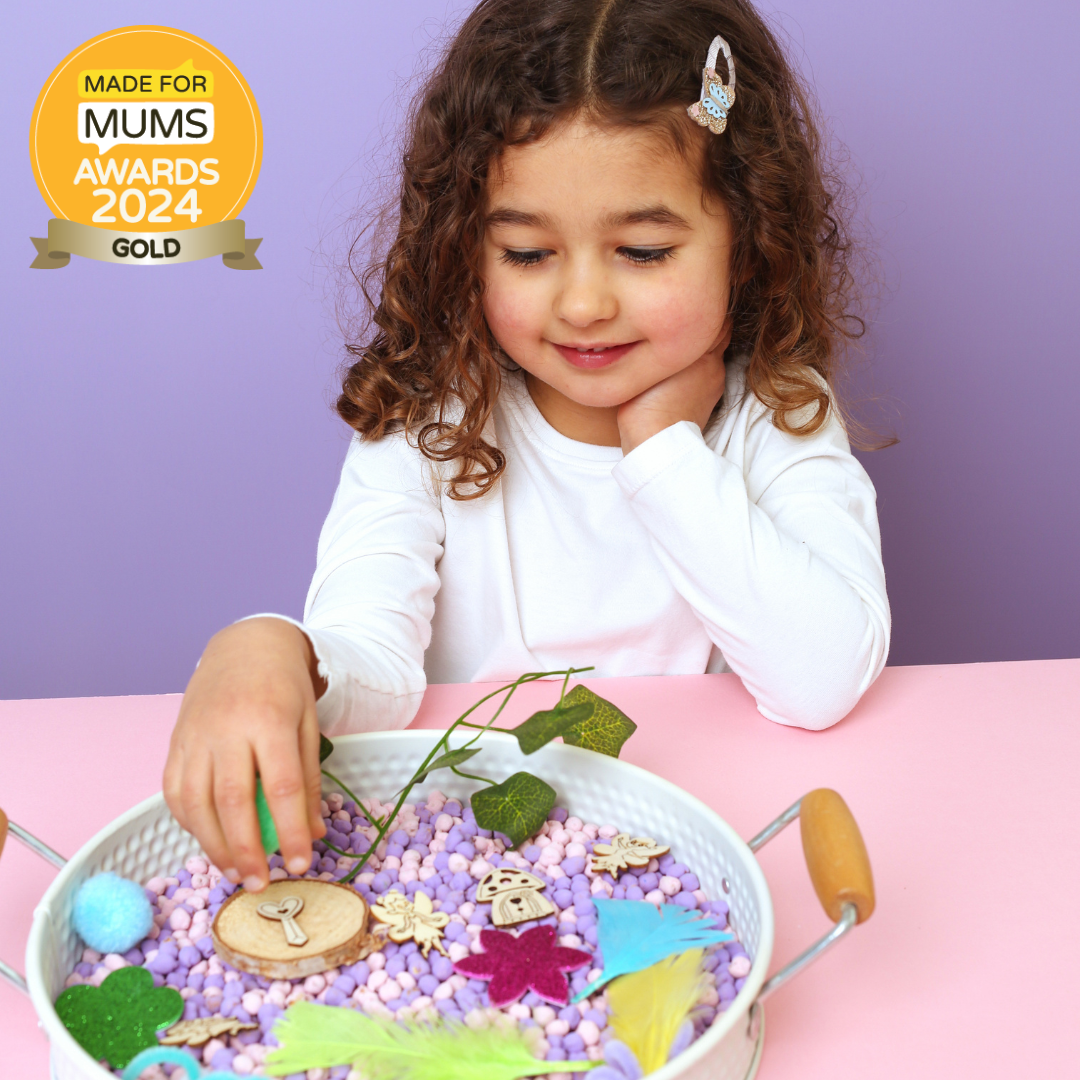 Child playing with a sensory bin filled with colorful materials on a pink table, with a gold 'Made for Mums Awards 2024 Gold' badge in the corner.