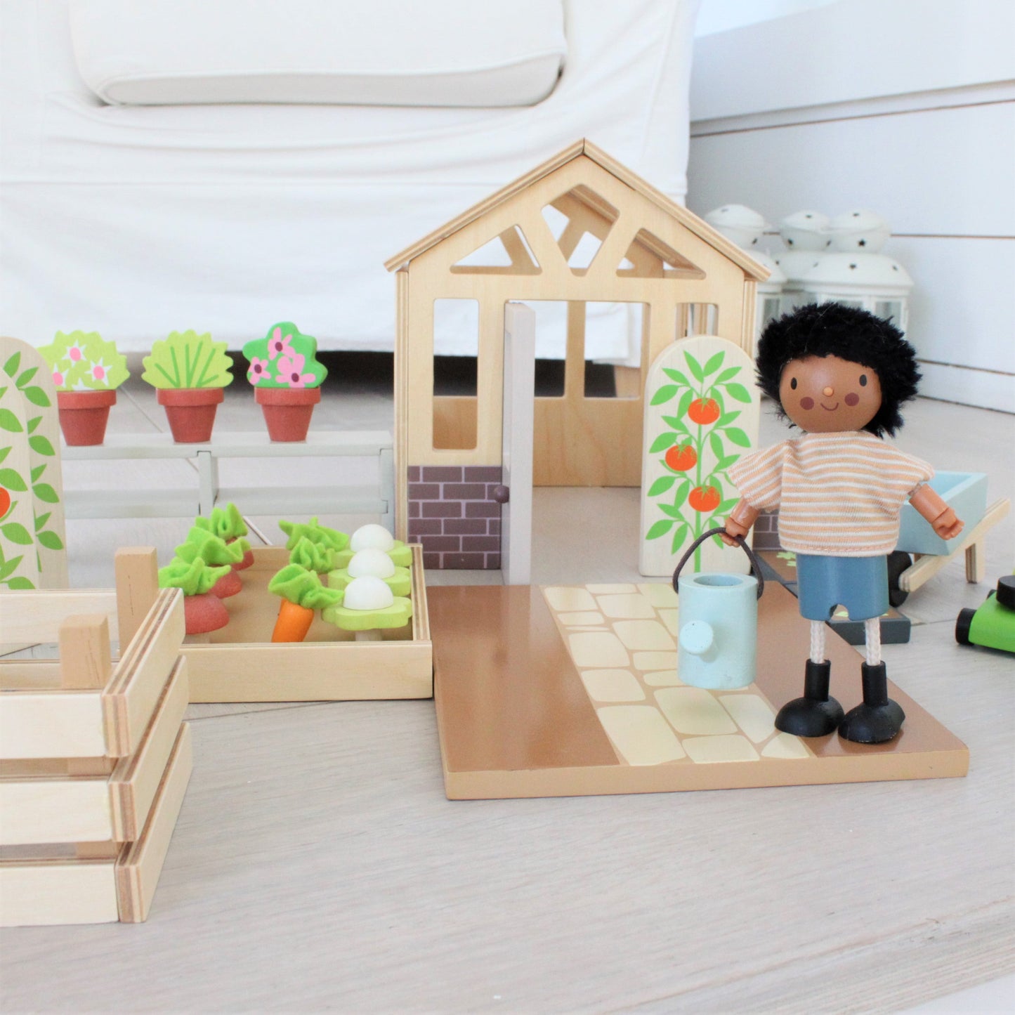 Wooden playhouse with garden and toy figure holding a watering can on a light wooden floor.