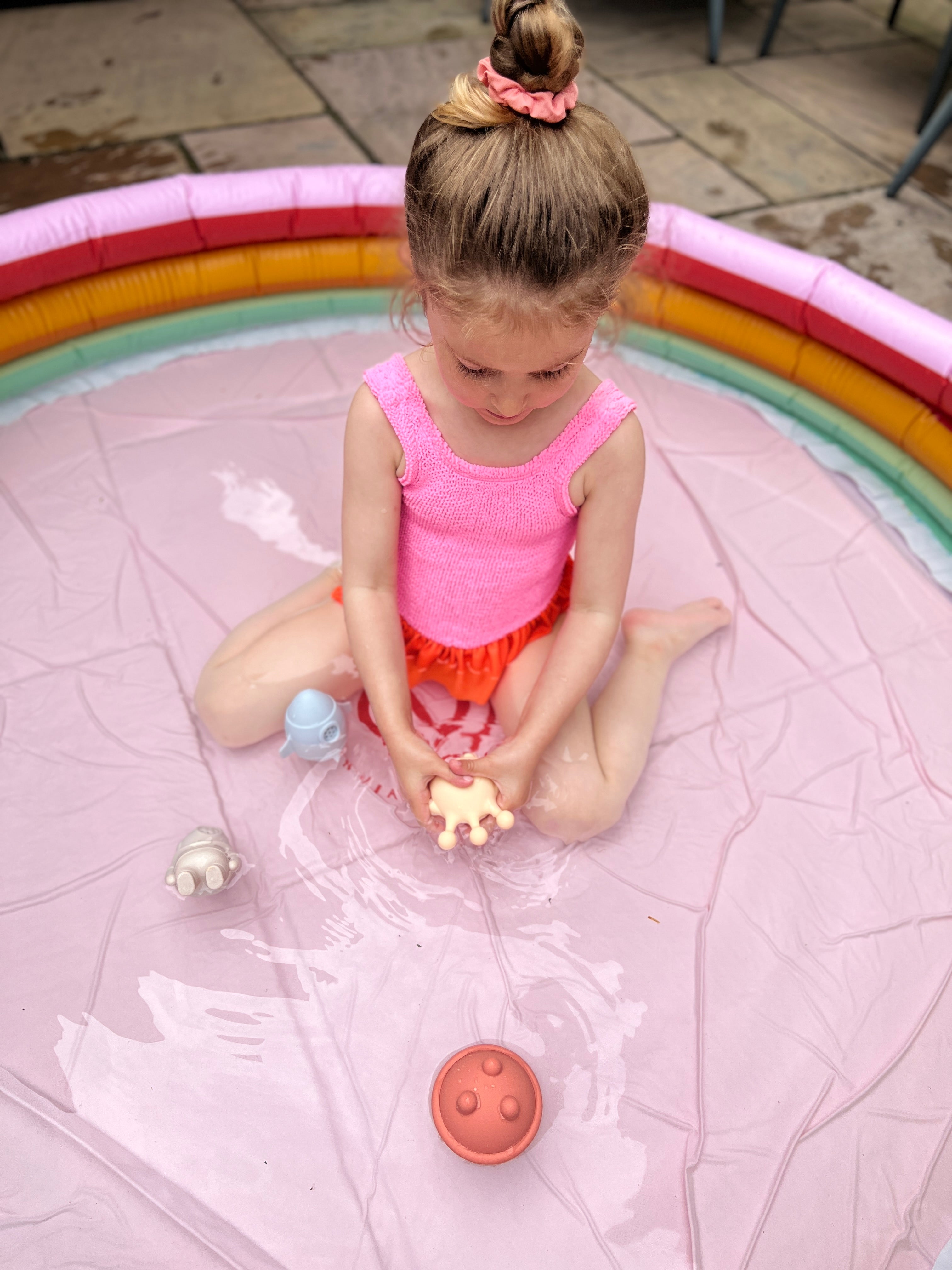 Child playing with space man bath toys toys in a small inflatable pool on a patio.