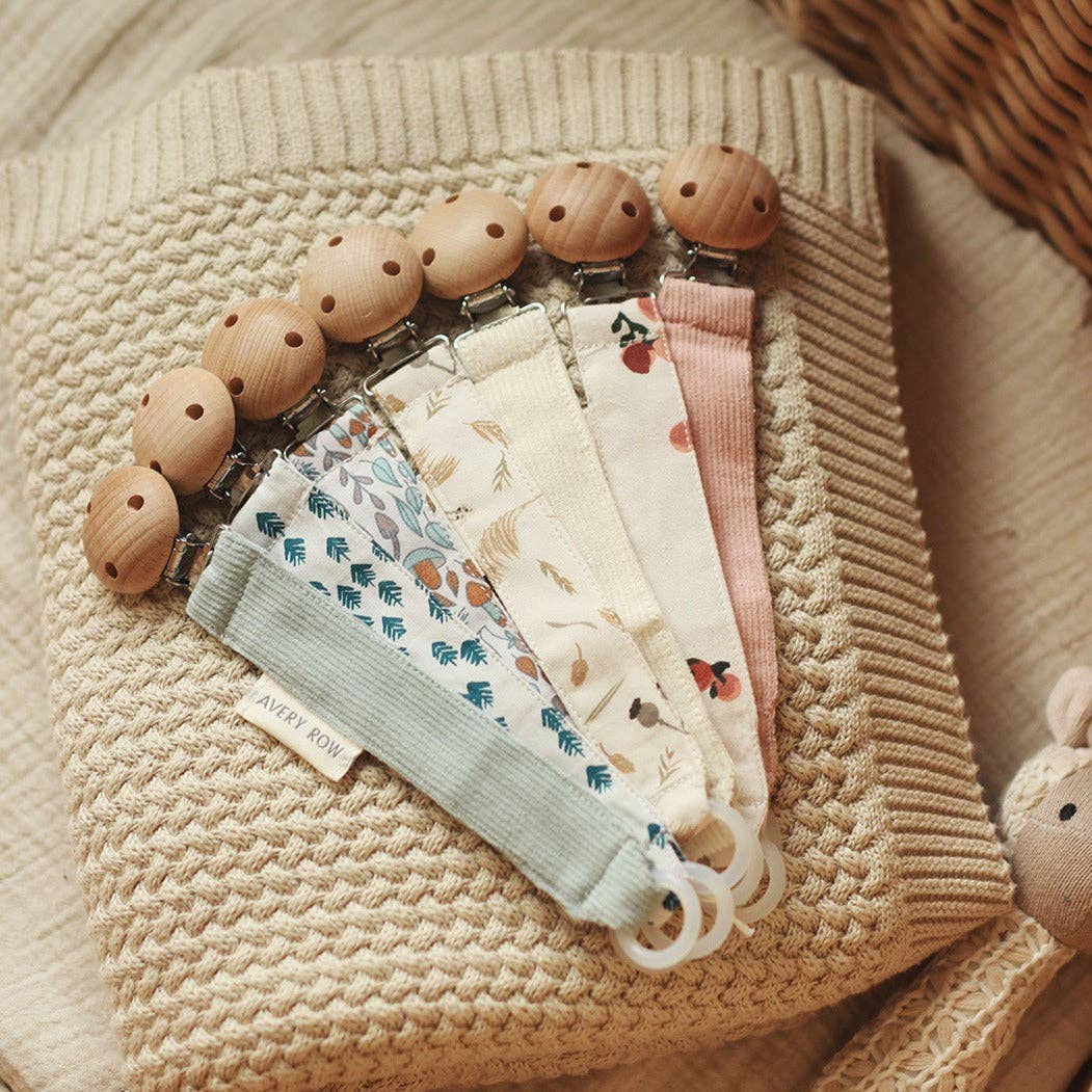 A fan arrangement of five fabric pacifier clips laid out on a textured beige knit blanket. Each clip features a rounded wooden clasp and a different strap design, including floral and botanical prints in soft, earthy tones—sage green, blue, cream, and dusty pink. A small fabric tag on one reads “AVERY ROW.” The clips end in silicone loops for attaching pacifiers. The cozy, neutral background enhances the natural, vintage-inspired aesthetic of the collection.