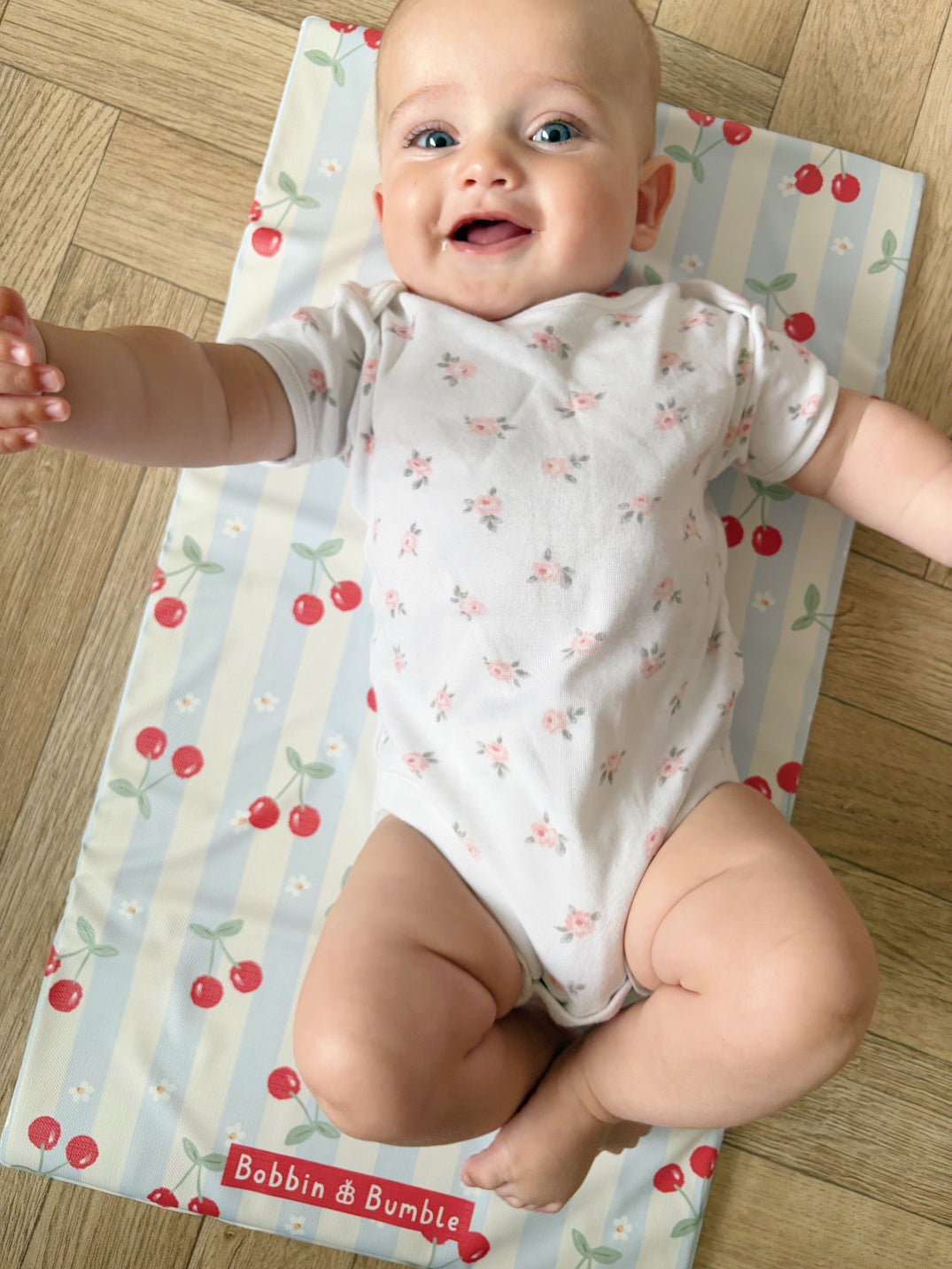 Baby in a floral onesie sitting on a colorful mat with 'Bobbin & Bumble' branding.
